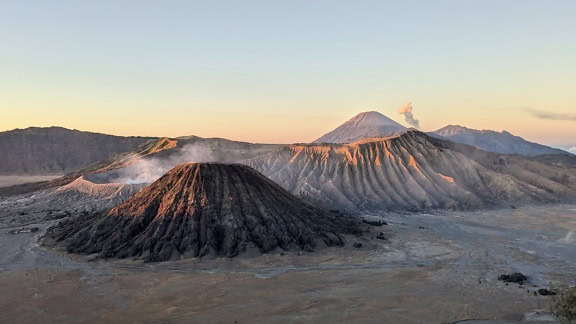 Una cadena montañosa con un volcán Bromo con humo saliendo de un cráter volcánico al atardecer