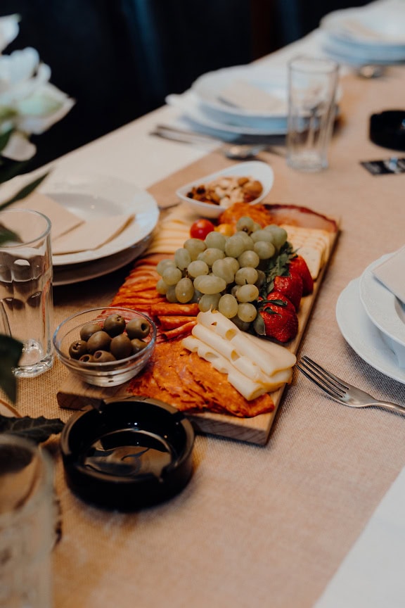 Ontbijttafel met borden en andere gerechten en eten erop, witte druiven met zelfgemaakte worst, olijf en harde kaas