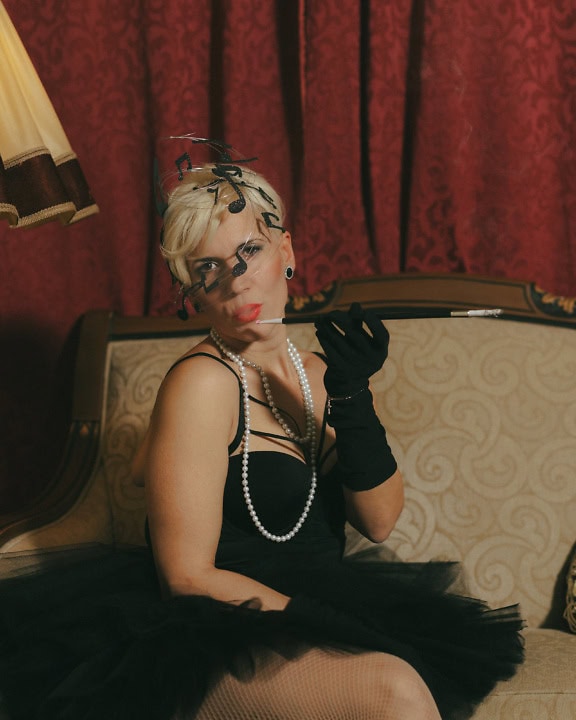 A woman sitting on the couch in a glamorous black dress and long gloves, wearing pearls and smoking a cigarette with a long cigarette holder