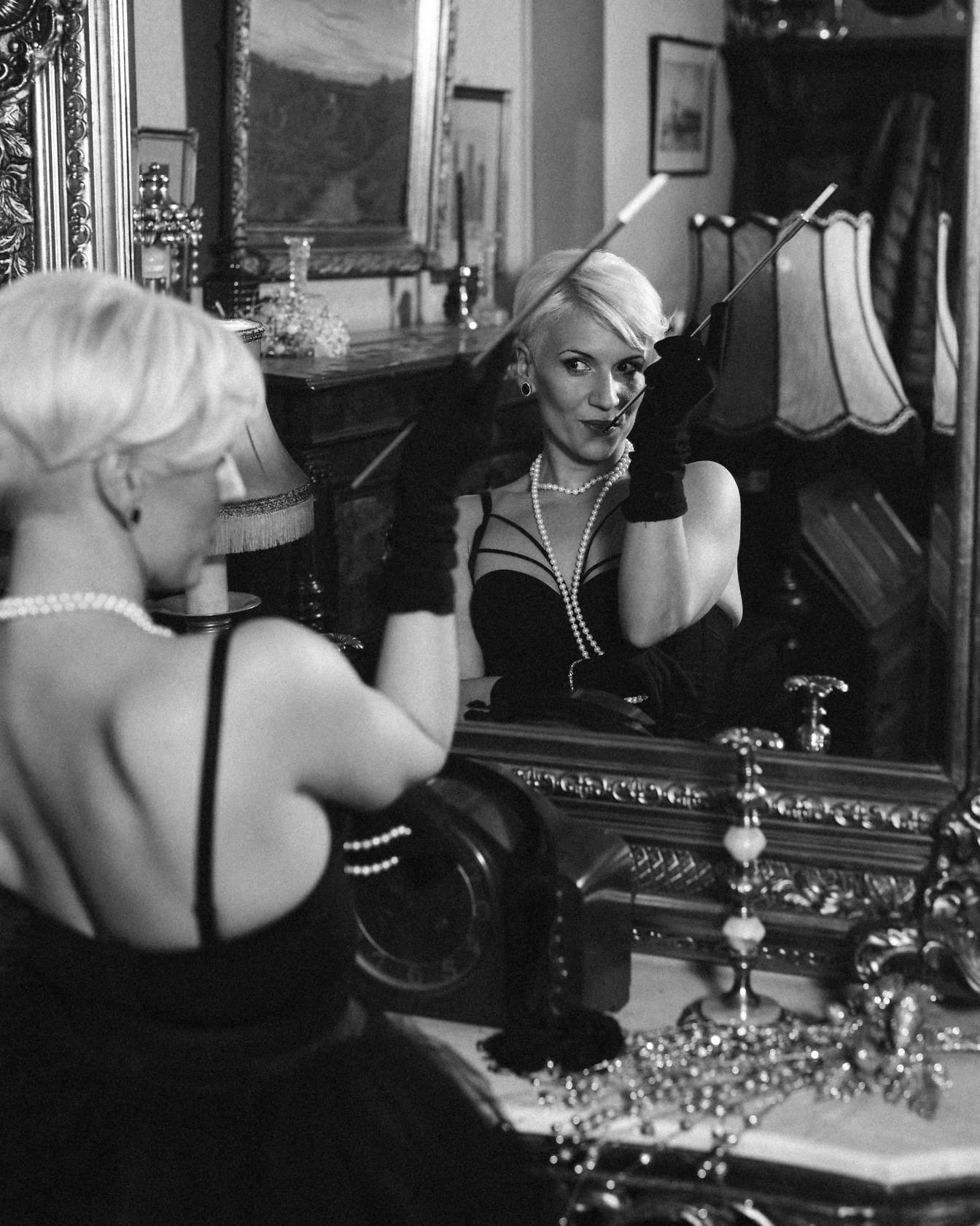 Black and white photo of a glamour woman posing in a black dress and gloves while holding a cigarette and looking in the mirror