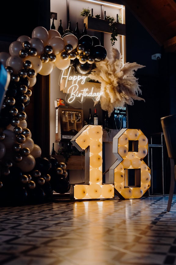 A lit up decoration in a shape of number 18 with balloons and a neon Happy birthday sign at the 18th birthday party, a celebration of an adult age