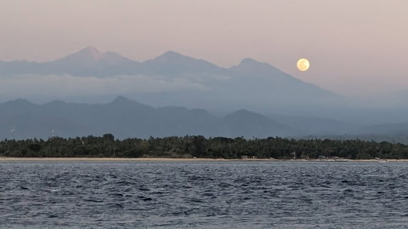 海の水平線に波が浮かび、曇りの山々に月が昇る海景のパノラマ