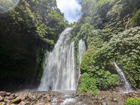 Persoon die op een rotsachtige rivierbedding onder een Tiu Kelep-waterval in bos van Indonesië loopt