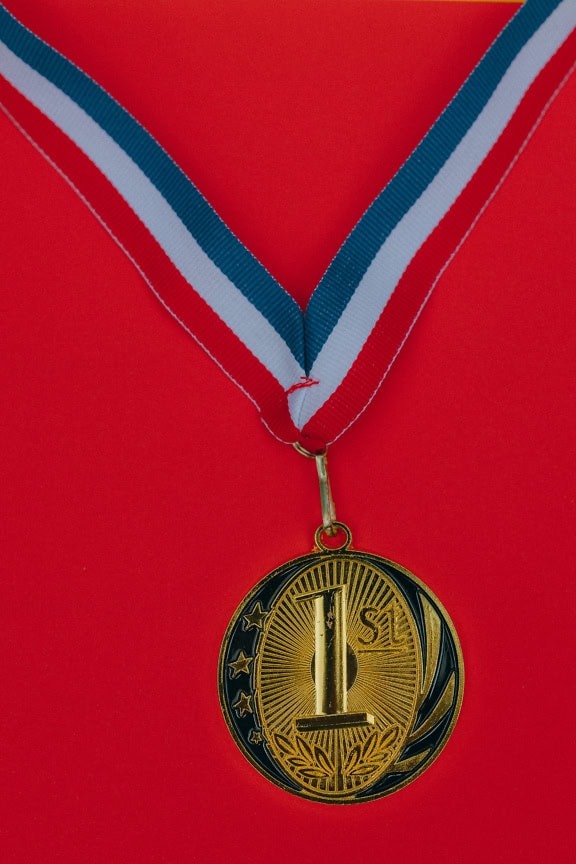 Gold medal with embossed first place emblem with red-white-blue ribbon on red background