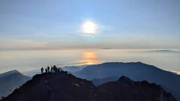 Panoramalandschaft bei Sonnenuntergang über einem nebligen Gebirge mit Silhouetten von Menschen auf dem Berg Rinjani, einem aktiven Vulkan in Indonesien