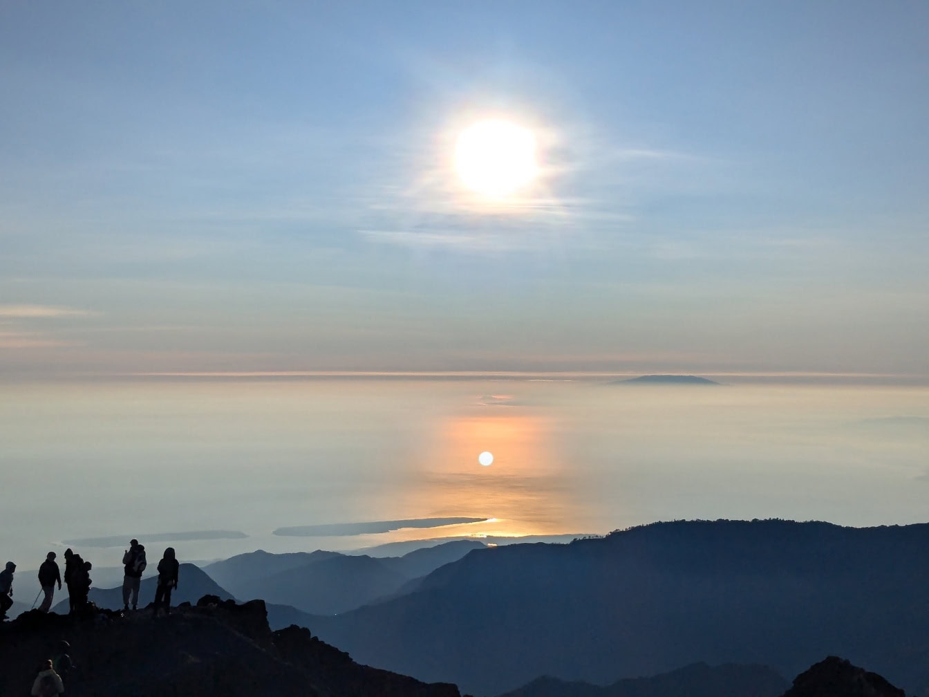 Skumring med glødende sol og silhuetter af mennesker, der står på et bjerg Rinjani, en aktiv vulkan i Lombok i Indonesien