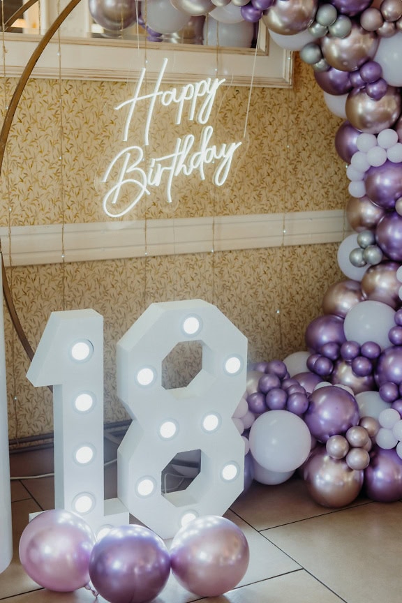 Large white number 18 and a neon Happy Birthday sign next to a large balloon arch with purplish-golden balloons