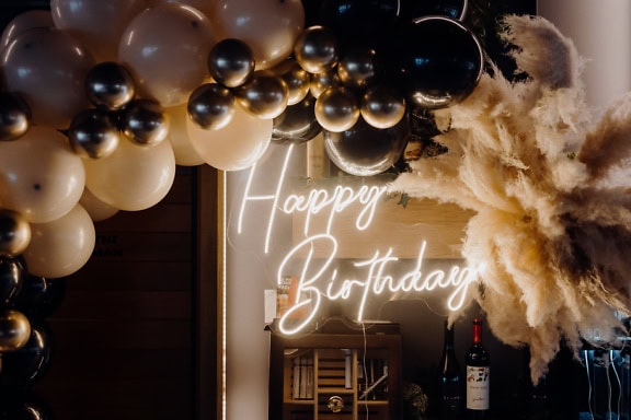 Hehkuva tyylikäs neon Happy Birthday -kyltti, jossa on kullanmustat ilmapallot ja pullo viiniä