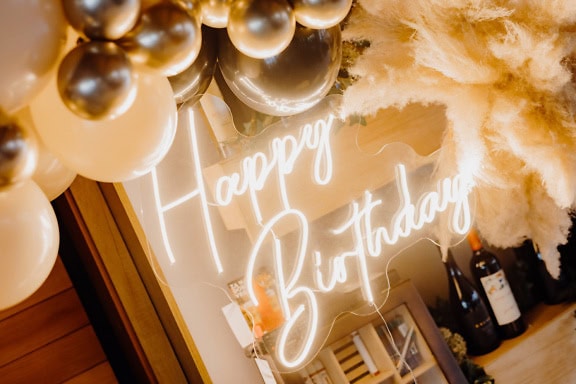 Hehkuva neon Happy Birthday -kyltti kiiltävillä kultaisilla ilmapalloilla