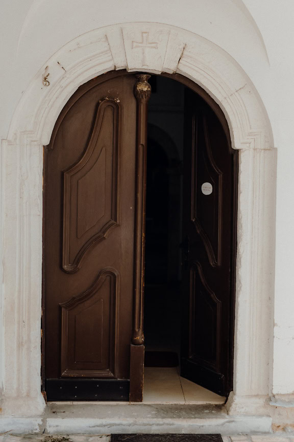 Um arco de pedra branca com uma cruz esculpida acima da porta de entrada marrom da igreja com uma porta aberta