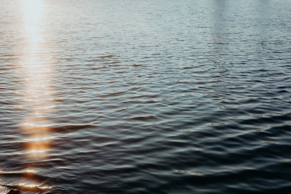 The surface of the water with calm waves, ripples and reflection of the sun’s rays as backlight