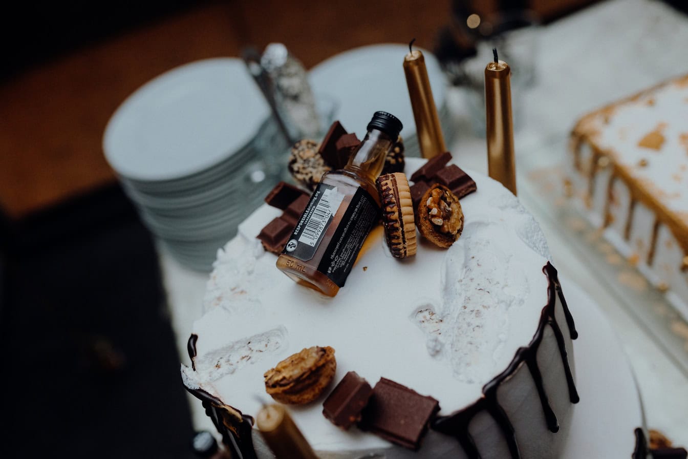 A white birthday chocolate cake decorated with melted milk chocolate, cookies and a small bottle of alcohol on top