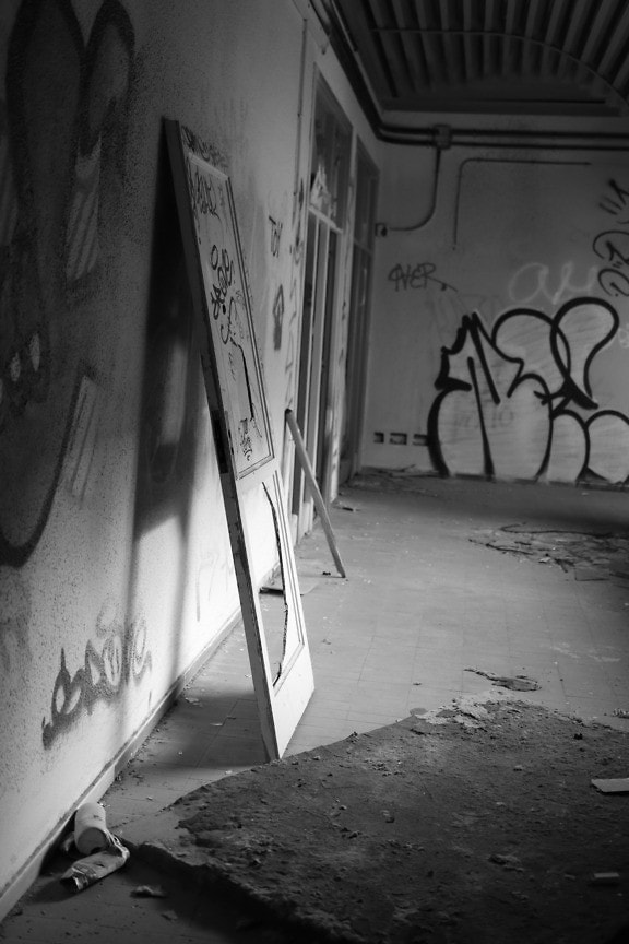 Black and white art photograph of an abandoned and ruined room by vandals with a broken door leaning against a wall with graffiti