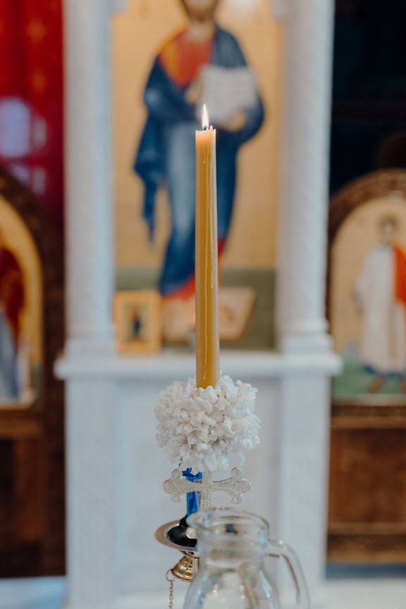 A flaming yellow candle with a cross on the candlestick and an icon of Jesus Christ in the background