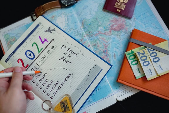 Hand holding a pen on a notebook with a map, passport, wristwatch and Euro banknotes, making a travel plan