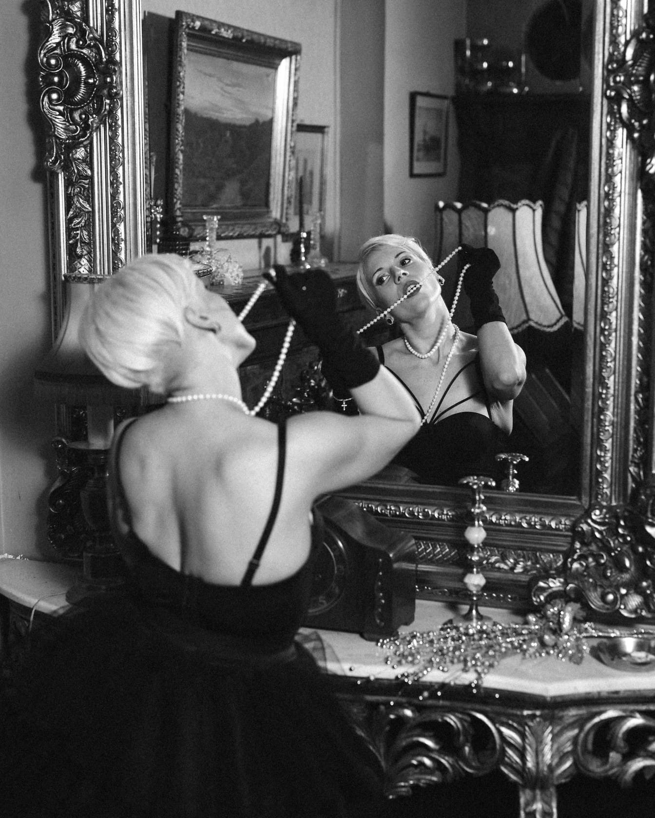 Seductive woman posing in a black dress and gloves holding a pearl necklace and looking at her portrait in the mirror