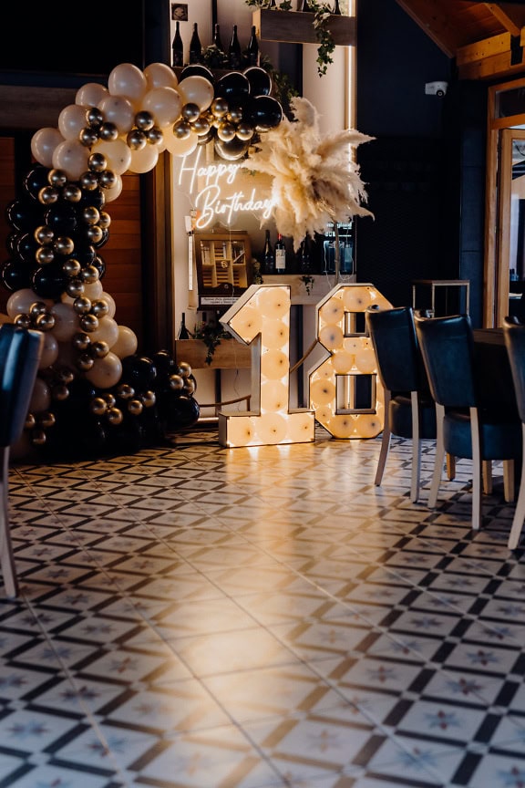 Una decoración de fiesta de cumpleaños dentro de un restaurante de vinos moderno-rústico, un cartel de Feliz Cumpleaños con globos junto a un número dieciocho (18)