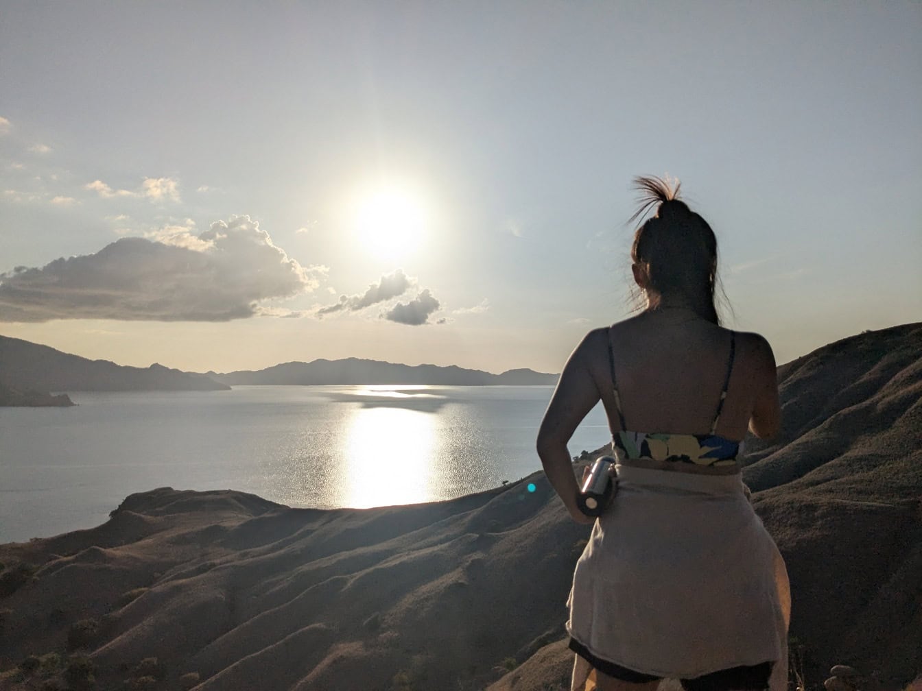 Mulher em pé em um ponto de vista no topo de uma colina com vista para uma baía com luz do sol brilhante como luz de fundo