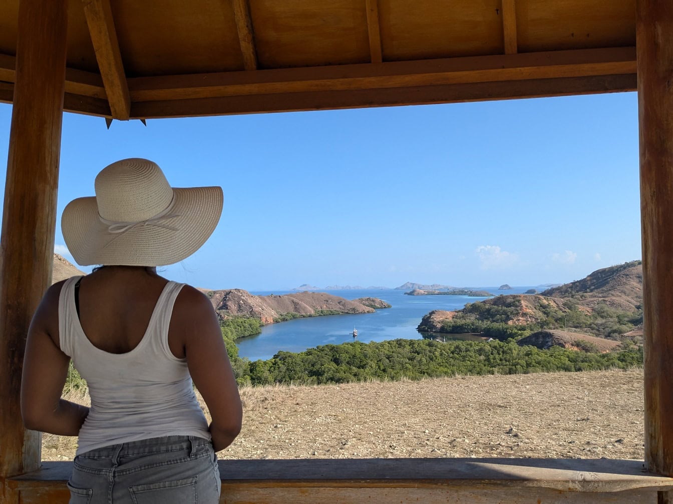 Donna con camicia bianca shouldrerless e cappello in piedi su un punto panoramico su una collina che domina una baia