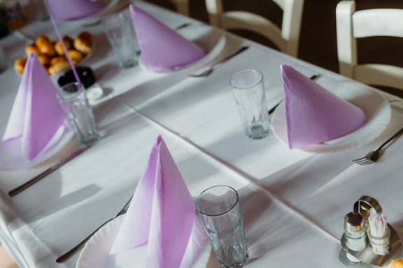 Close-up of a dining table with plates with pink-purple paper napkin and with glasses and other cutlery