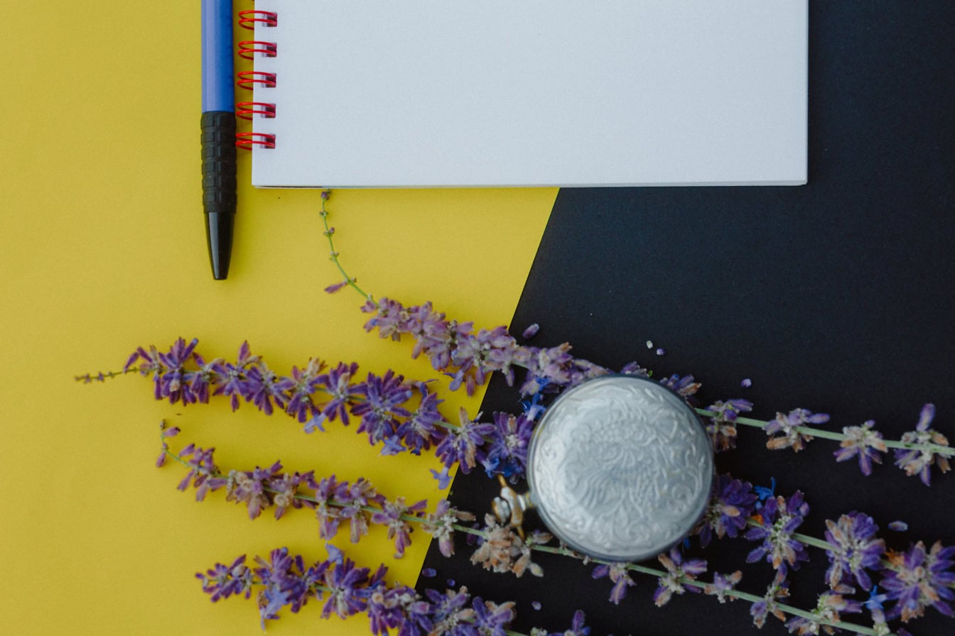 Buku catatan dan pena dengan bunga lavender dan arloji saku analog pada permukaan kuning dan hitam