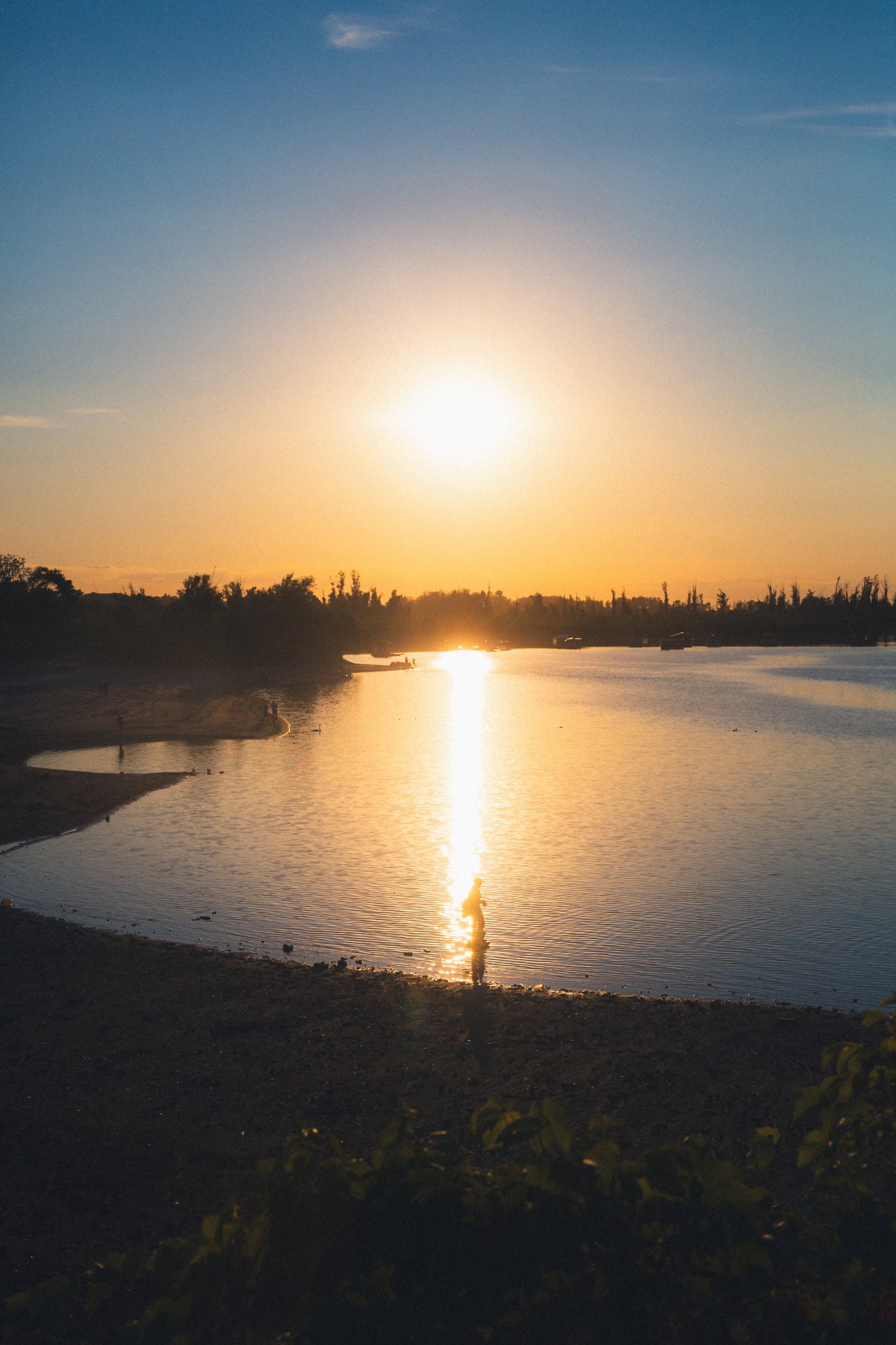 O siluetă a unei persoane care stă în apă puțin adâncă lângă malul unui lac la apus