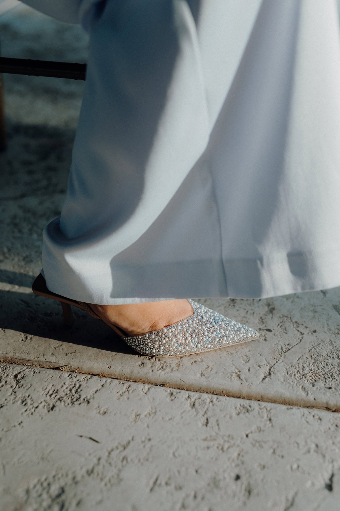 El pie de una persona con un vestido blanco y una sandalia de tacón alto con decoraciones de circonitas