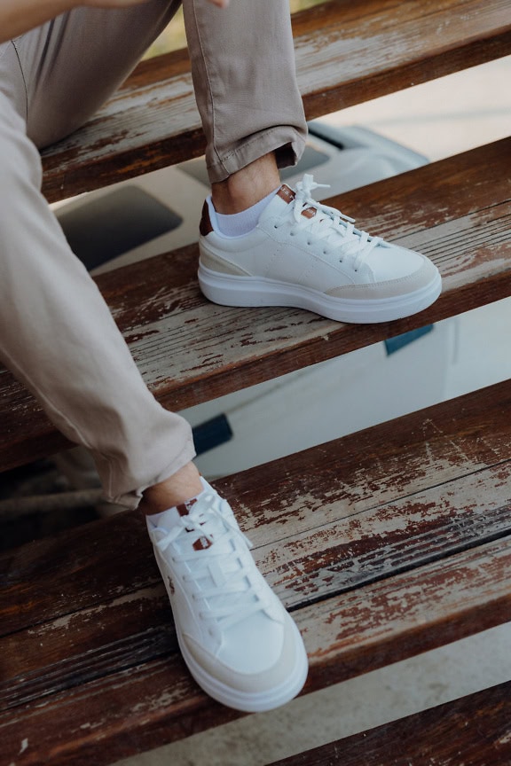 Piernas de hombre con pantalón y zapatos-zapatillas deportivas blancas en una escalera de madera