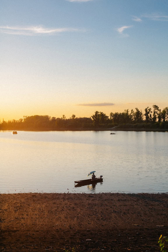 Henkilö kalastaa veneessä sateenvarjon alla järvellä pari venettä vedessä auringonnousun aikaan