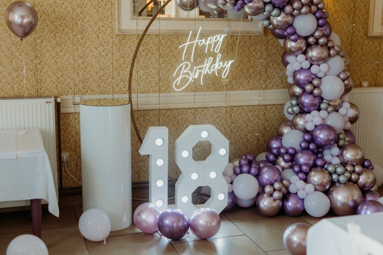 Large white and purple balloon arch with a lit up neon Happy Birthday sign and a decoration in a shape of number eighteen (18)