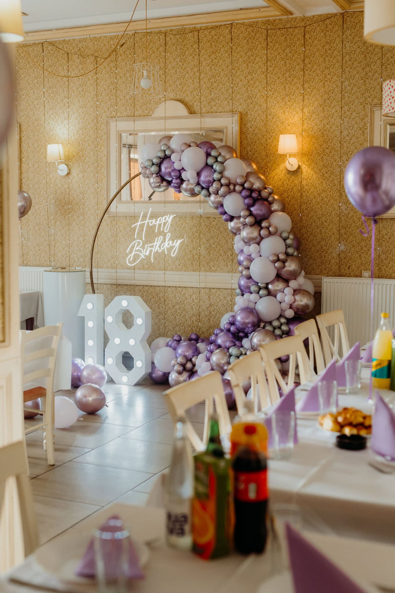 Room decorated for birthday party with white and purplish balloons and a lit up neon Happy Birthday sign