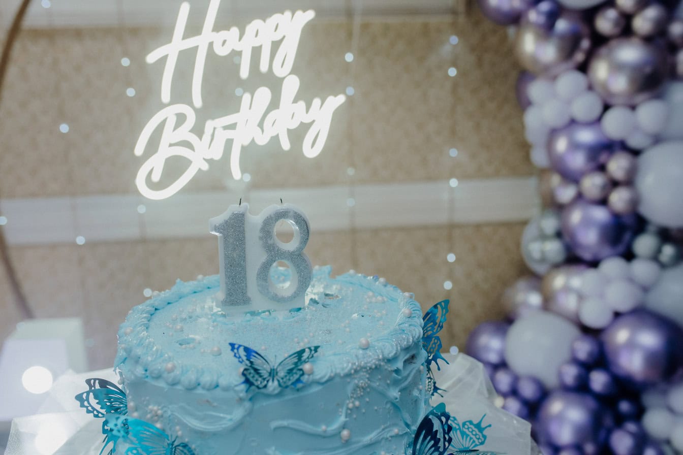 Birthday cake with blue icing, a candle in the shape of the number 18 on the top and a lit up white neon Happy Birthday sign