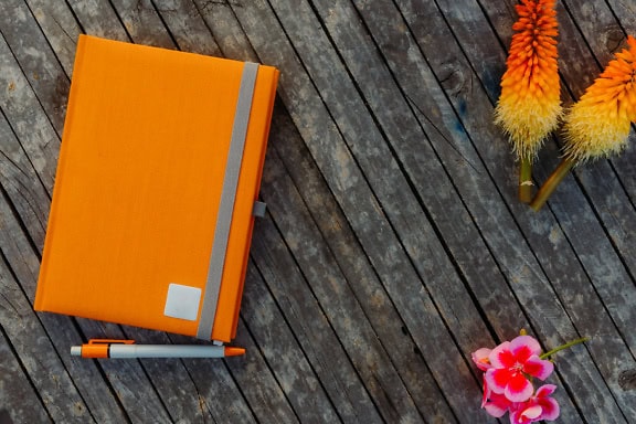 Orange notebook and pencil with orange-yellow flowers on a wood surface