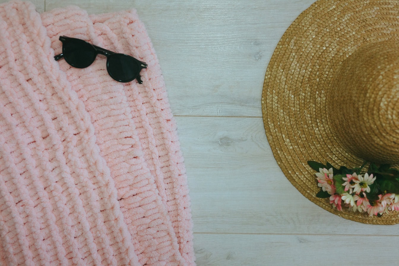 Pull en tricot rose avec des lunettes de soleil et un chapeau de paille sur une surface en bois blanc