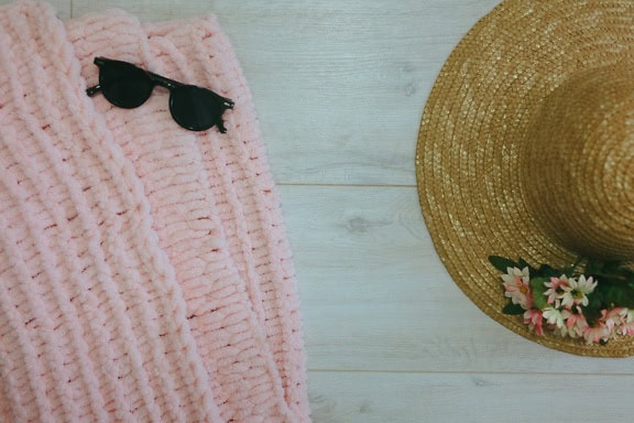 Pink knitted sweater with sunglasses and a straw hat on a white wooden surface