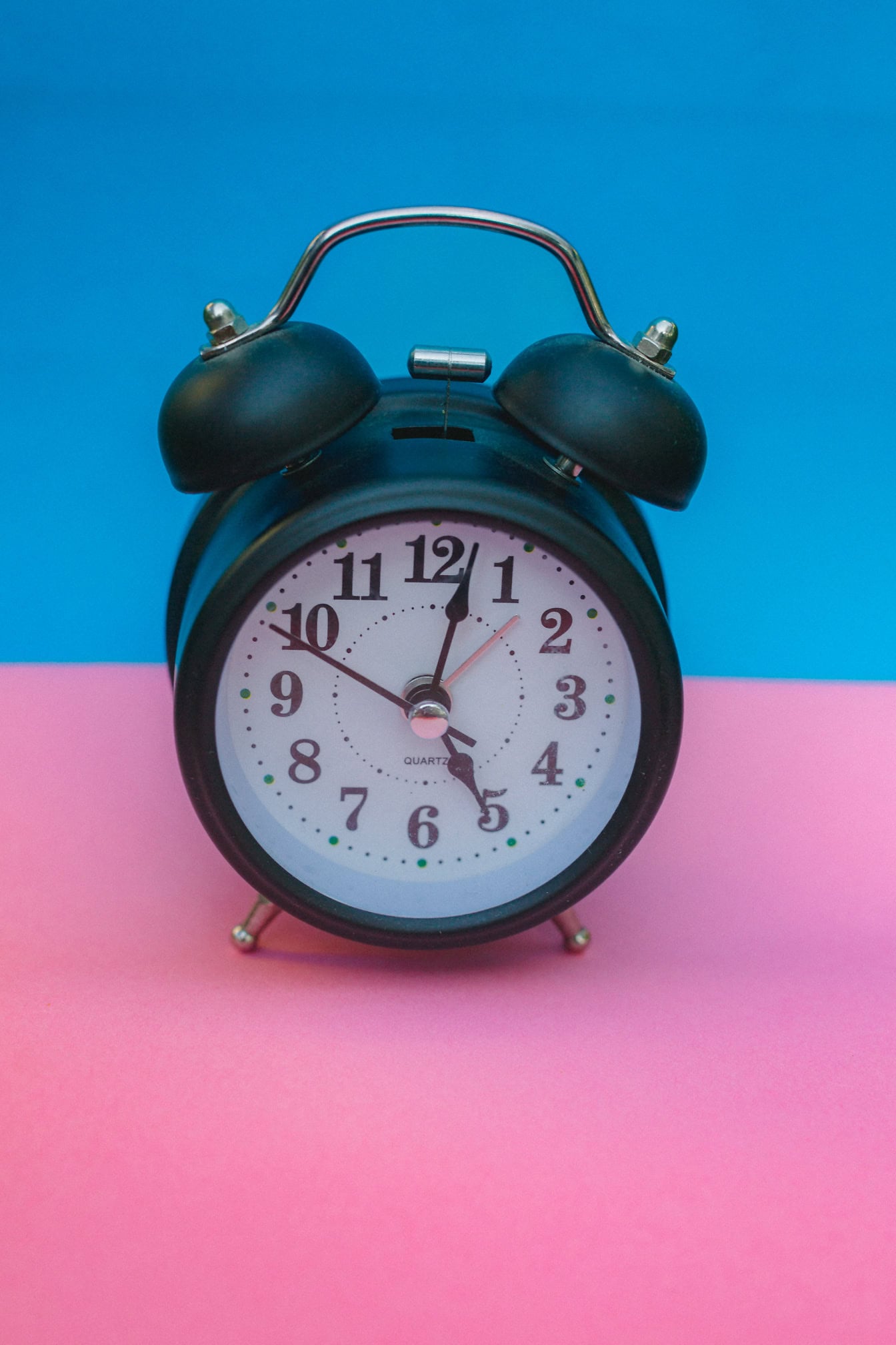 Black analogue alarm clock with wake-up bells on a pink and blue background