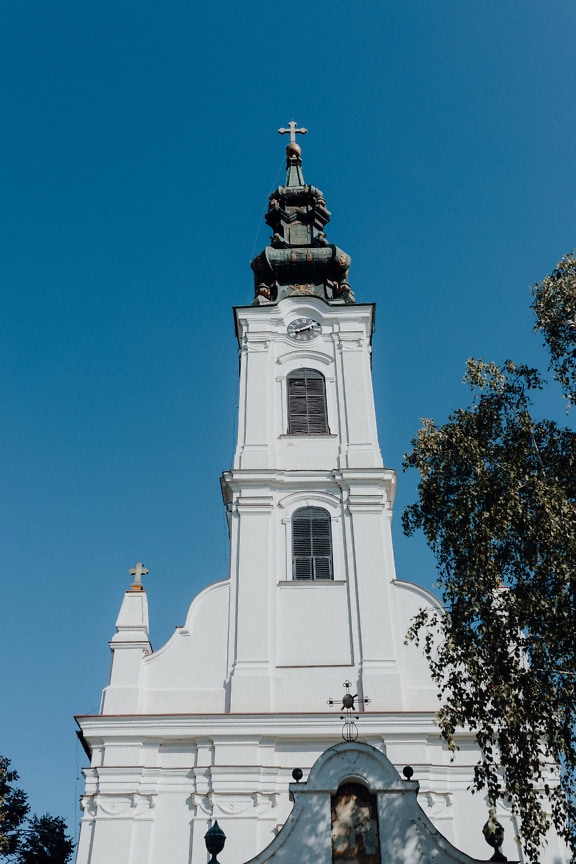 Hvidt klokketårn med et ur og et kors fra den serbisk-ortodokse kirke for Johannes Døberens fødsel, Backa Palanka, Serbien