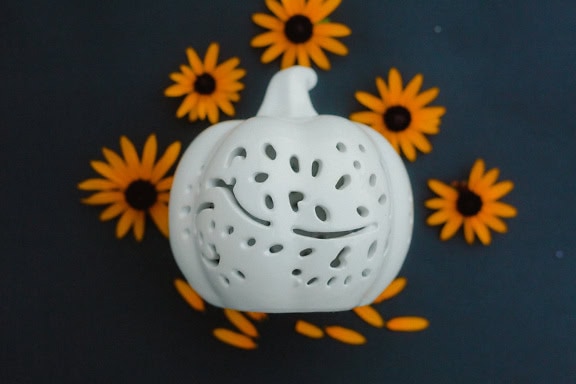 Ceramic figurine of a white pumpkin with carved holes surrounded by yellowish flowers on a black background