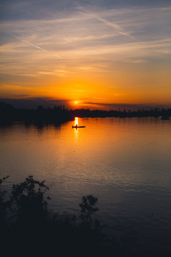 Vertikales Landschaftsfoto einer Silhouette eines kleinen Bootes auf einem See bei Sonnenaufgang mit orange-gelbem Himmel im Gegenlicht