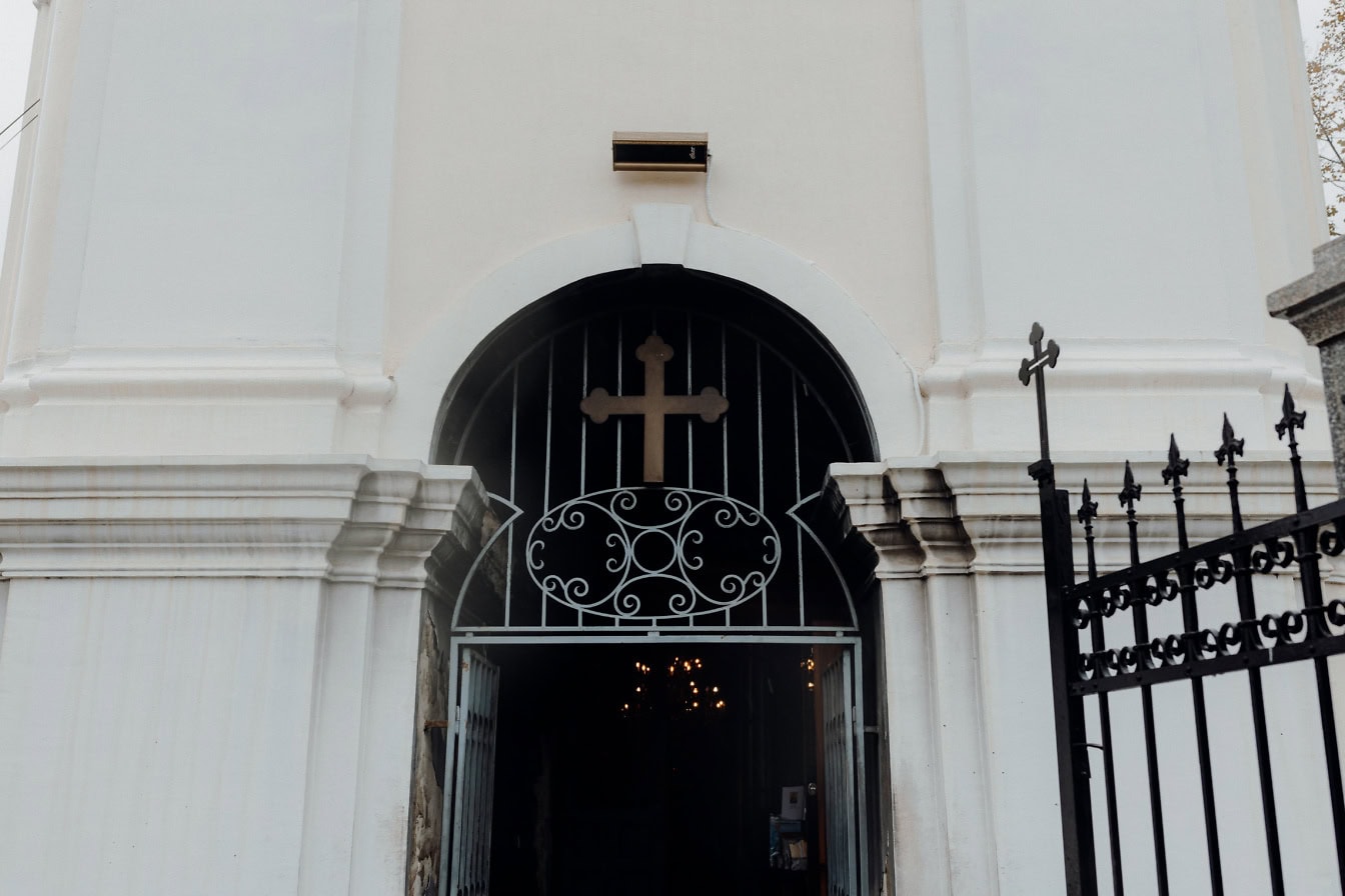 Cast iron gate with Christian cross above at the dark entrance of white church