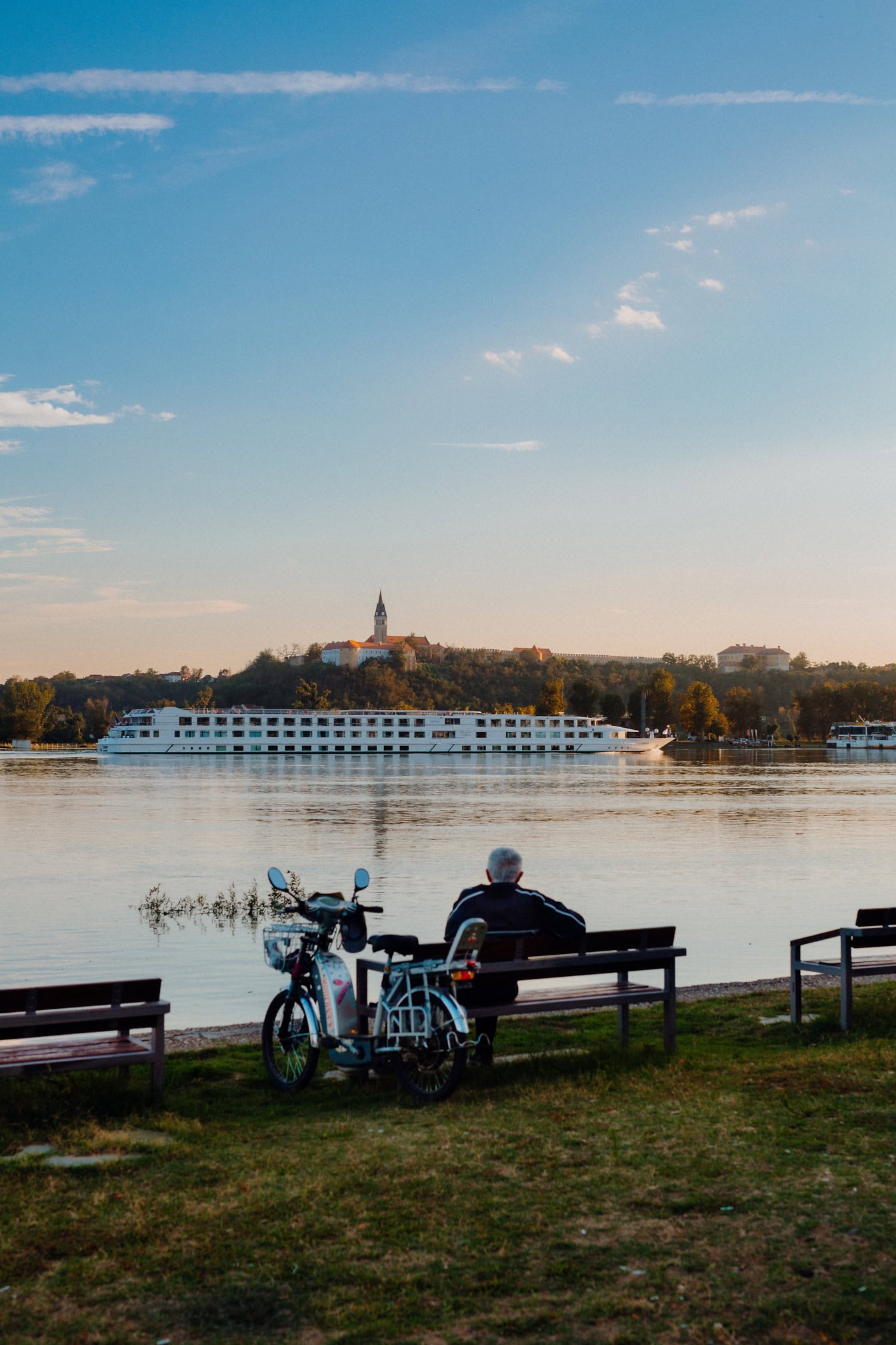 En mand, der sidder på en bænk med knallert ved siden af en bænk og nyder udsigten over Donau-floden med et stort hvidt krydstogtskib, der sejler