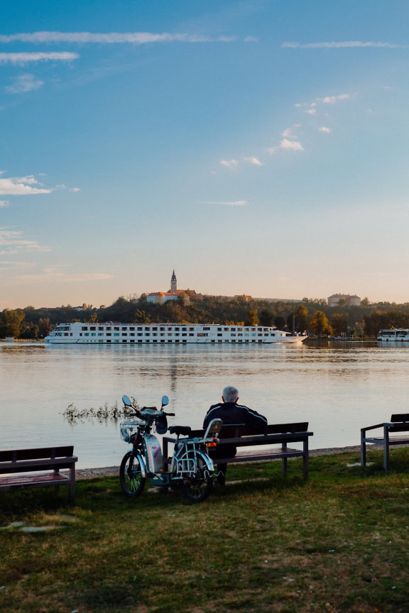 Un bărbat care stă pe o bancă cu un moped lângă o bancă și se bucură de priveliștea fluviului Dunărea cu un vas mare de croazieră alb care navighează