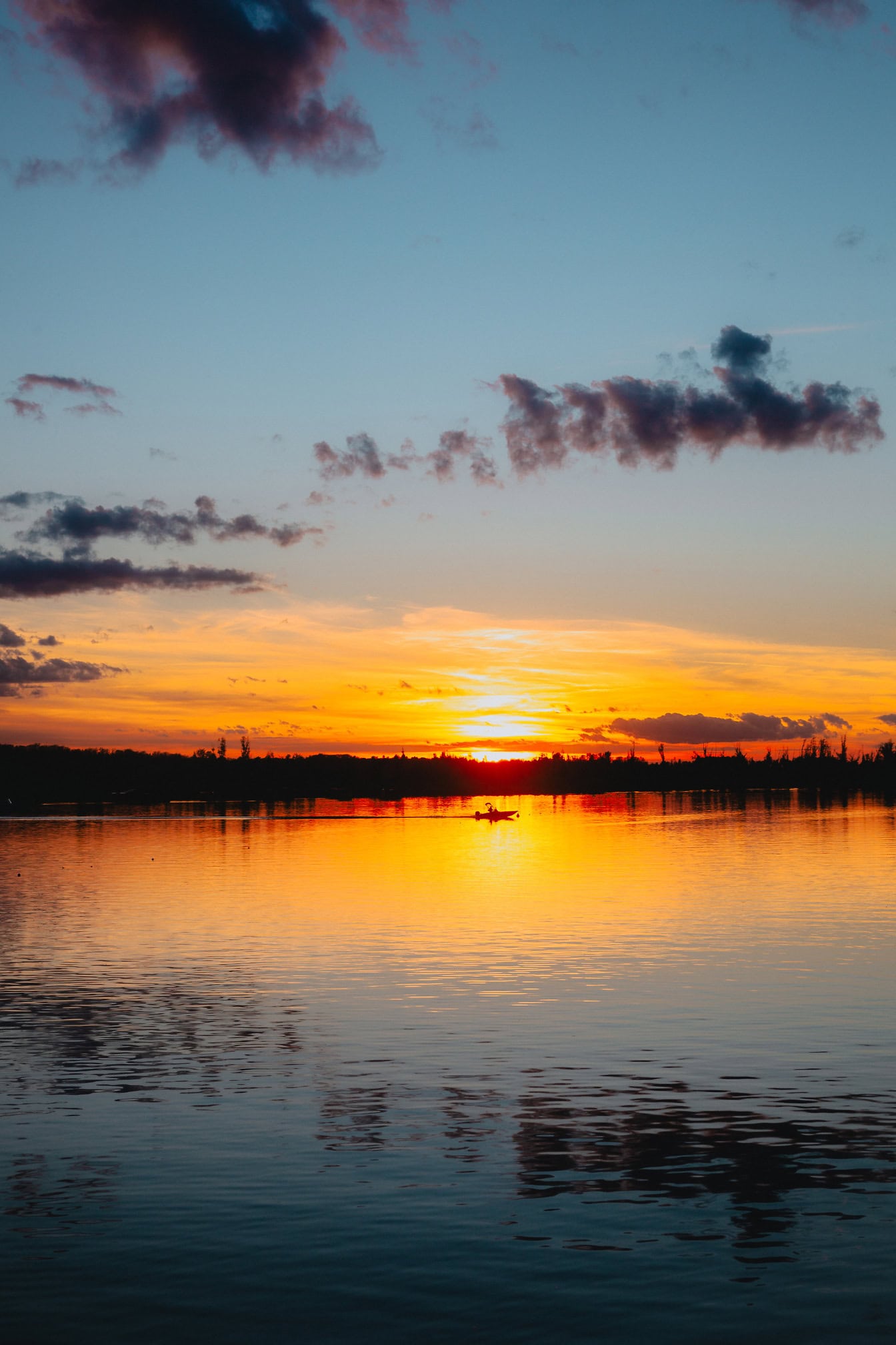 Apus de soare pe malul unui lac cu reflexia luminii solare galben-portocalii pe apă