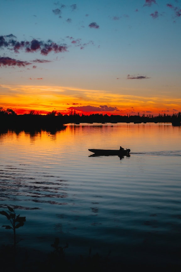 Silhuet af en lille båd med en levende orangerød himmel, der reflekteres på vandet ved solnedgang i Tikvara Naturpark, Bačka Palanka