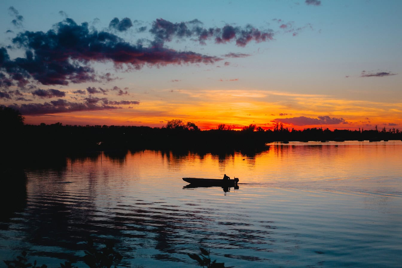 Puesta del sol dramática con luz solar vibrante en el cielo sobre la orilla del lago con la silueta de la persona en el bote en el agua