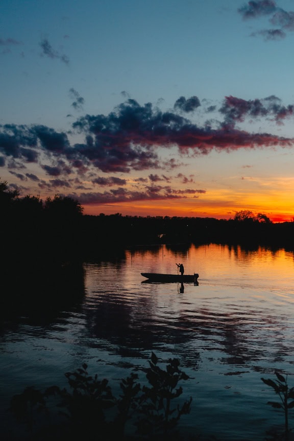 Kontur av en fiskare i en båt på en sjö vid mörk sen solnedgång