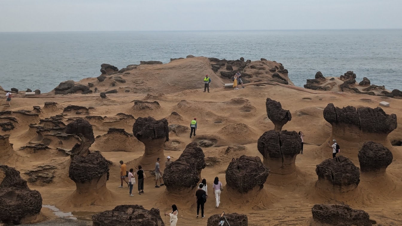 Turistacsoport áll egy homokos területen az óceán mellett, furcsa sziklaképződményekkel a Yehliu geoparkban, egy fokon a Wanli kerületben, Új-Tajpejben, Tajvanon