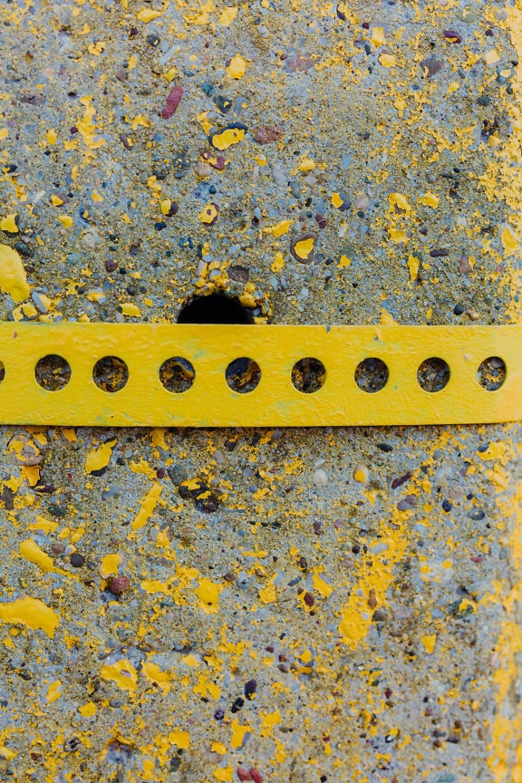 A yellow metal strip with holes on concrete painted with yellowish paint that is peeling, and