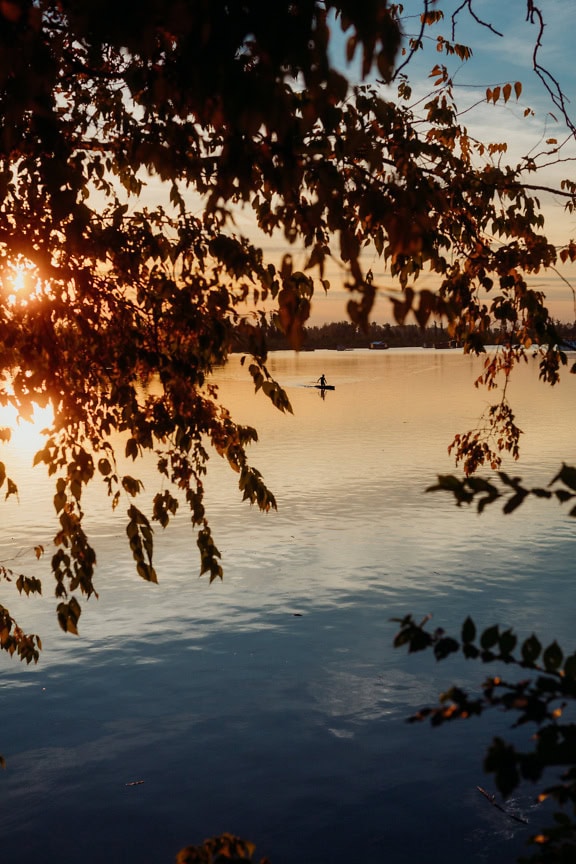 Kanootissa olevan henkilön siluetti järvellä, jossa kirkkaat auringonsäteet ovat taustavalona auringonlaskun aikaan