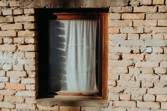 Ein Fenster mit weißem Vorhang auf einer unverputzten Backsteinwand